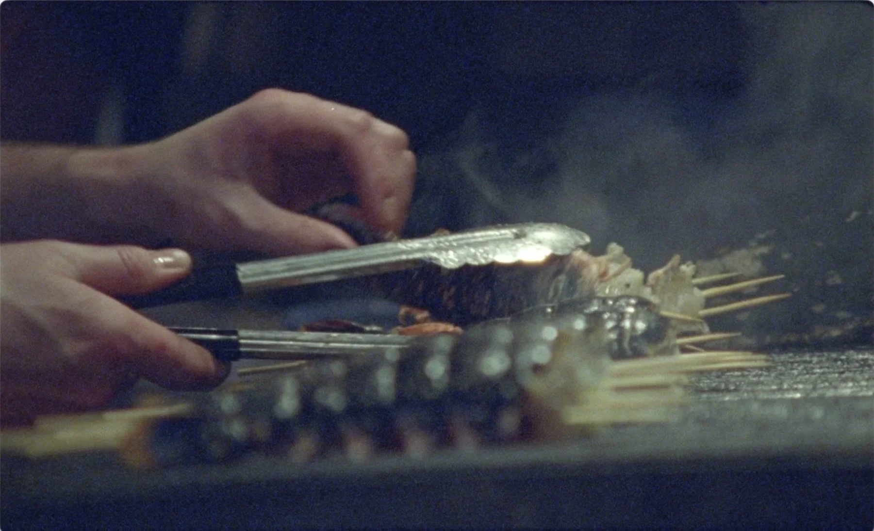 Hands working tongs over wood grill with skewers and smoke rising at Mountain
restaurant Soho, 16mm film James Dougan