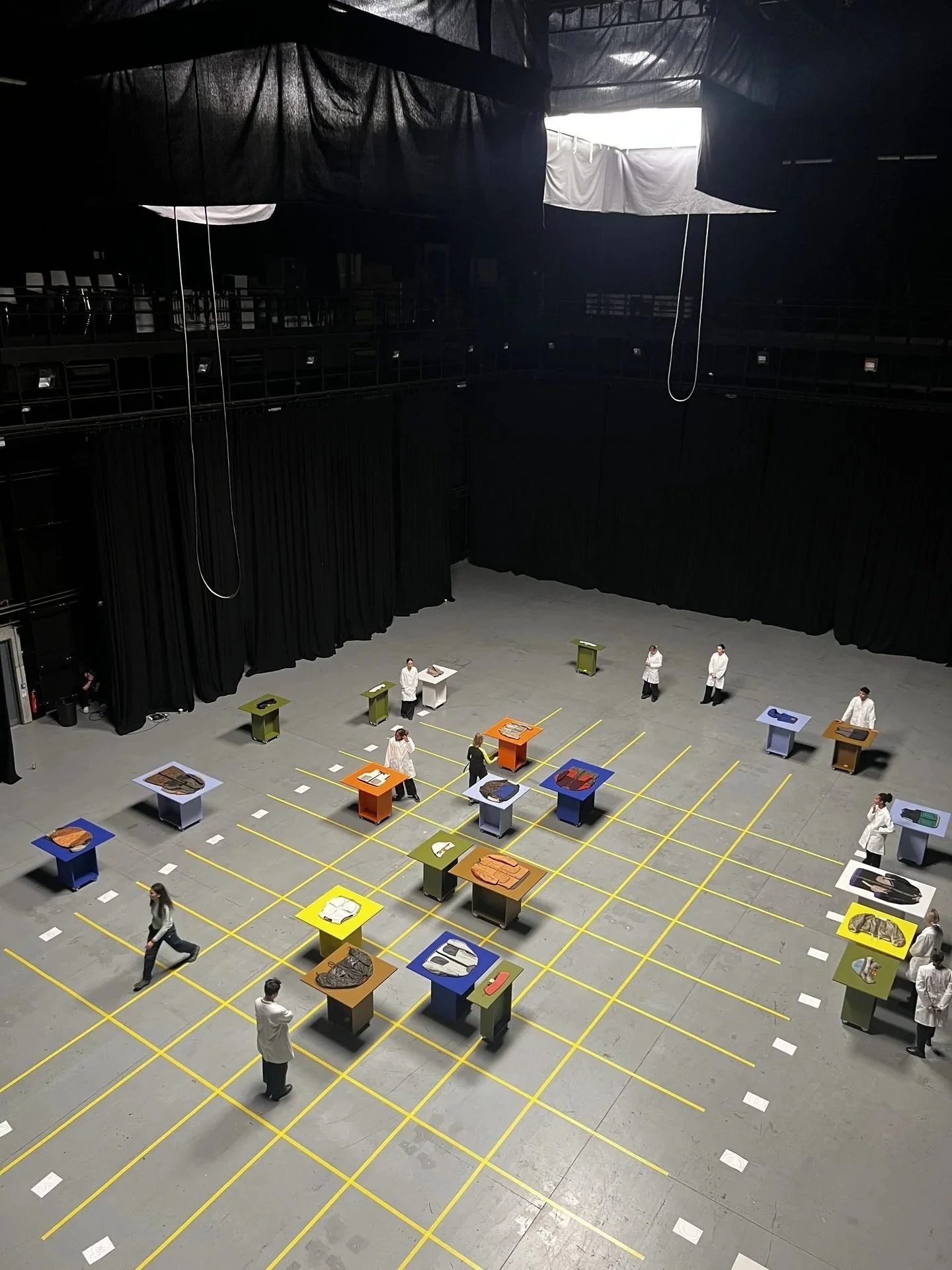 Overhead walkway view of performers placing Marfa Stance garments on colour-blocked
plinths in grid, DOP James Dougan