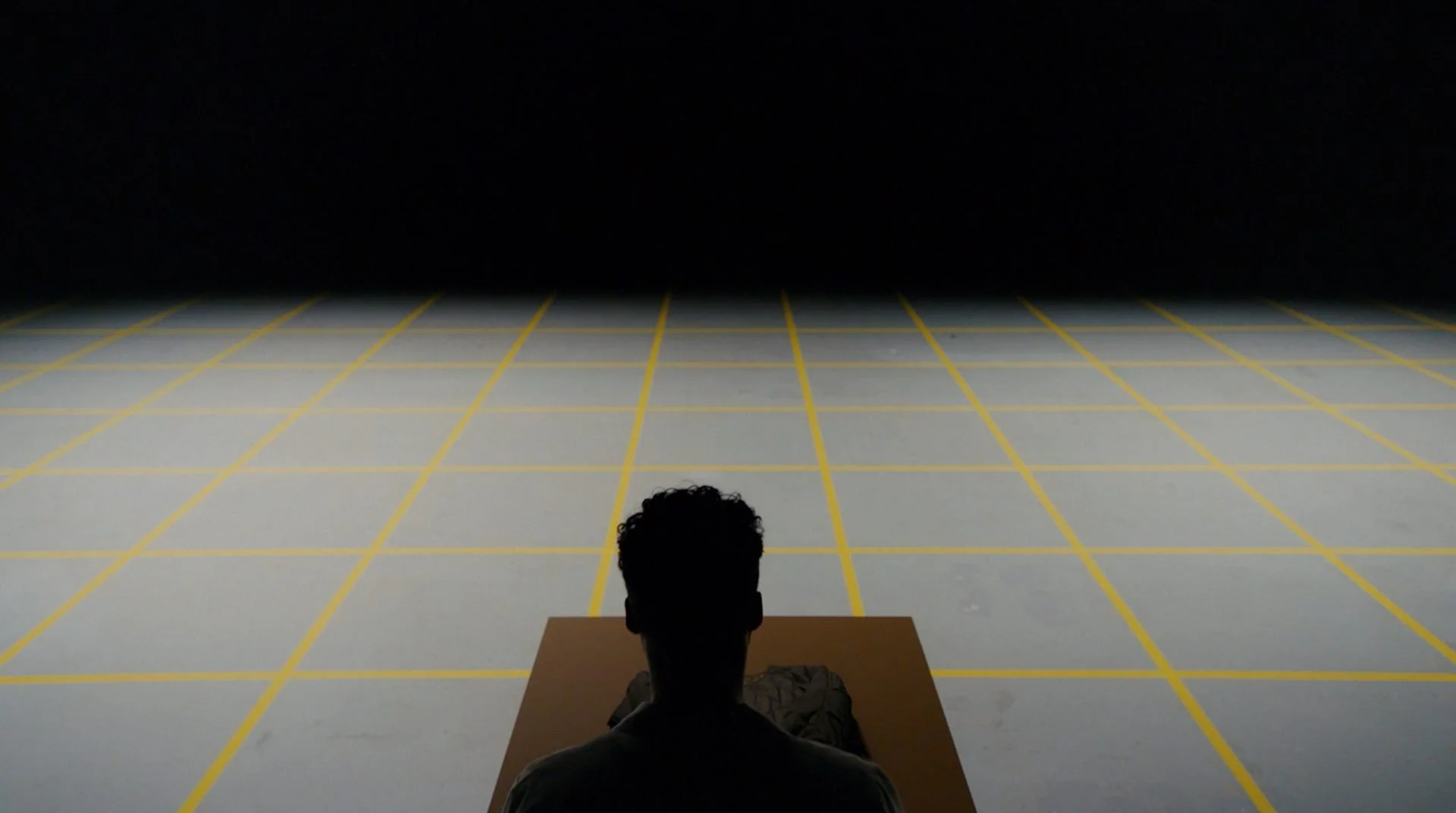 Marfa Stance film still: silhouetted figure seated at plinth on lit yellow-gridded floor, overhead
angle, DOP James Dougan