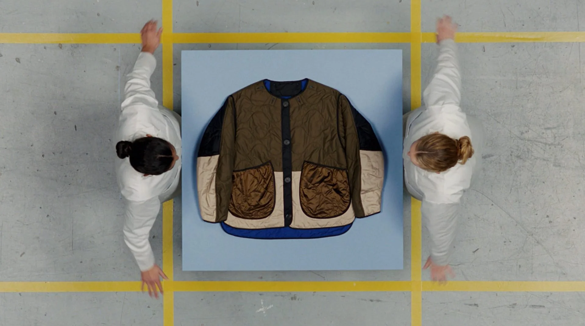 Marfa Stance film still: two performers symmetrically placing quilted jacket onto blue plinth,
overhead, DOP James Dougan