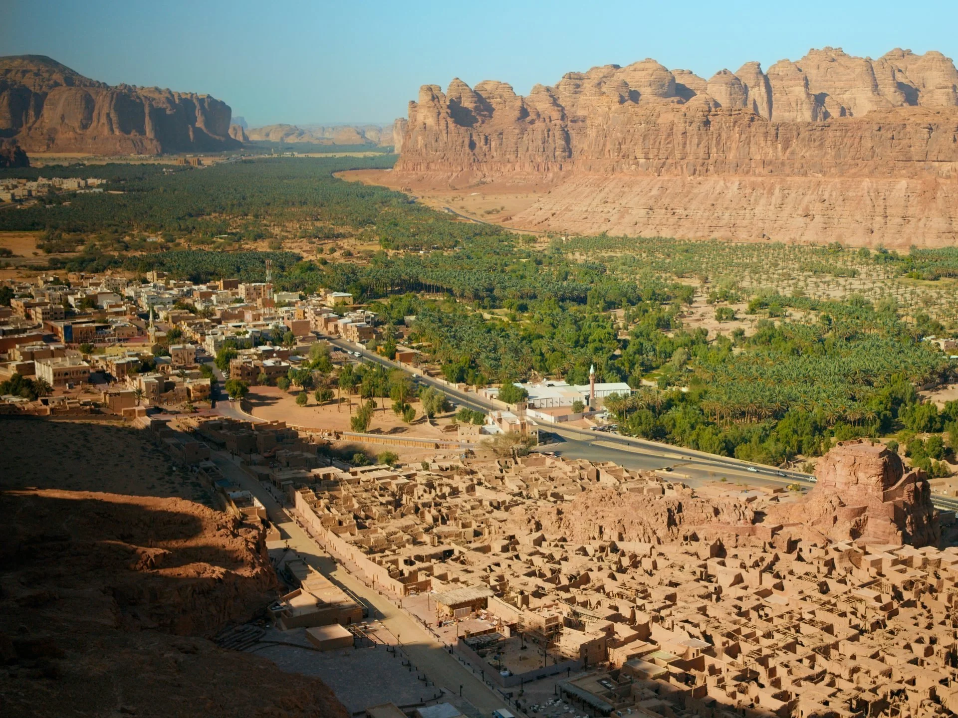 Wide drone shot of AlUla - the ancient mud-brick old town ruins in the foreground, the
green oasis valley running through the middle, and towering sandstone cliffs rising
beyond