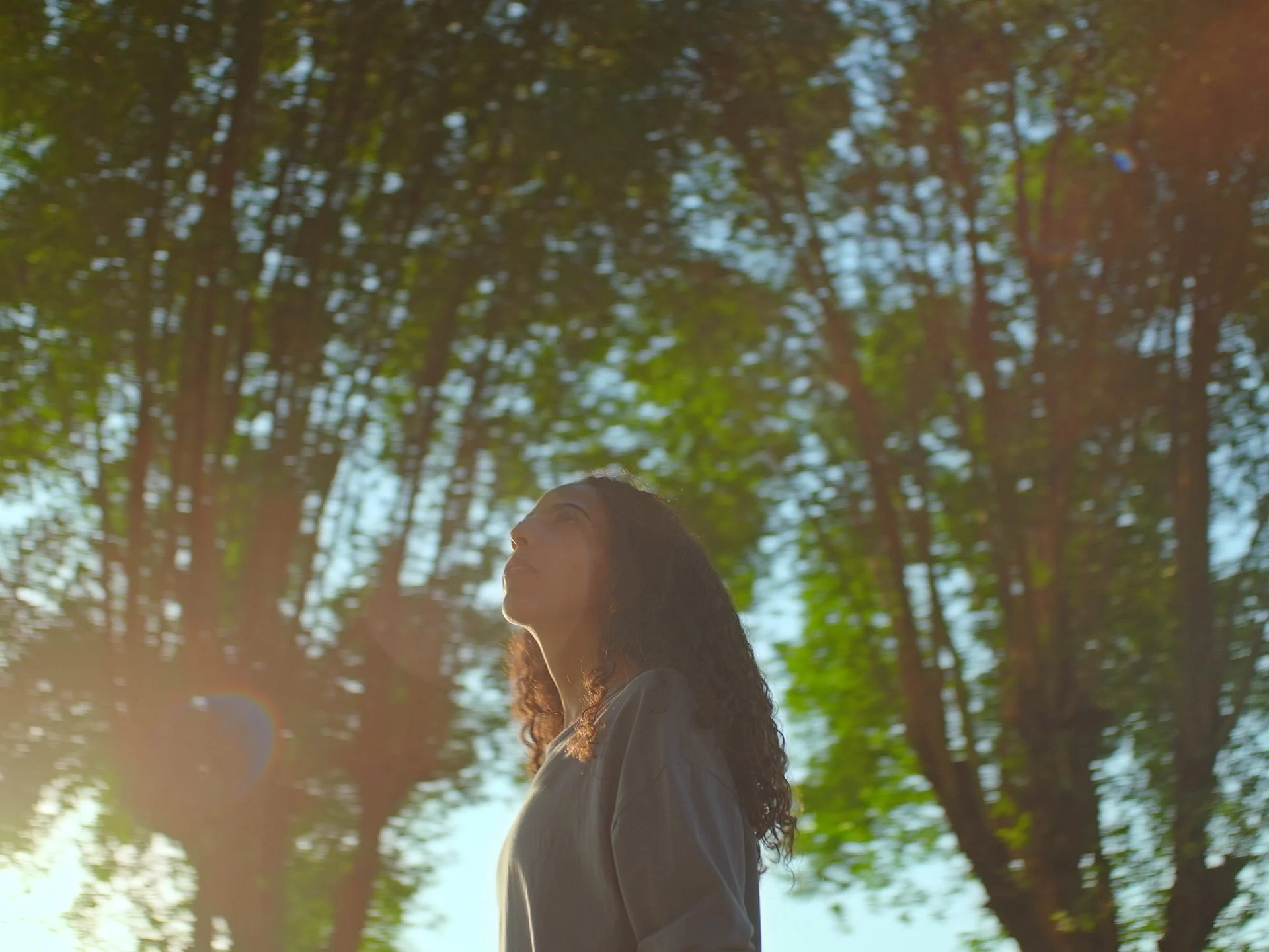 A young Saudi woman looks upward with eyes closed, surrounded by tall trees with
warm sunlight filtering through - filmed at King Salman Park, Riyadh, for Green Cities