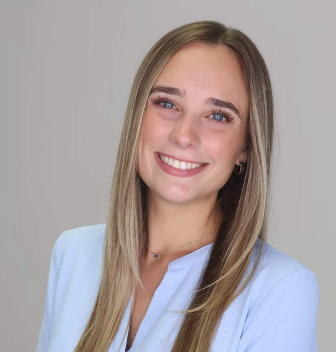 Portrait of a young woman with long blonde hair, blue eyes, smiling, wearing a light blue blazer and small hoop earrings, against a plain light gray background.