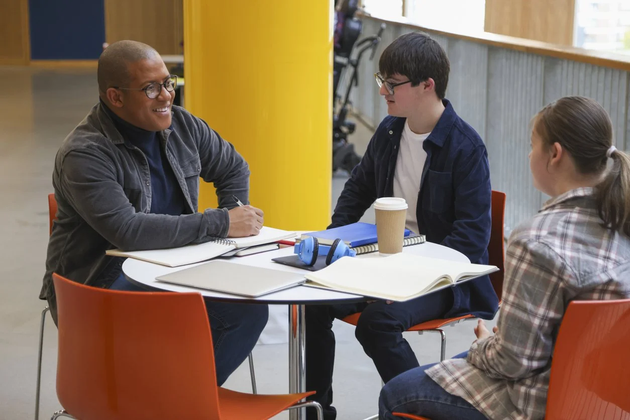 Three people sitting at a round table having a discussion in a brightly lit indoor setting, with notebooks, a coffee cup, and headphones on the table.