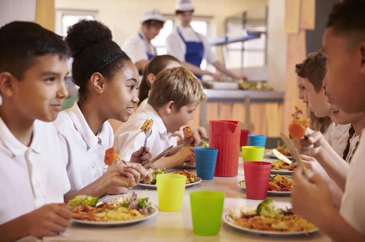 Children sitting at a table in a school cafeteria eating lunch while two school staff members prepare food in the background.