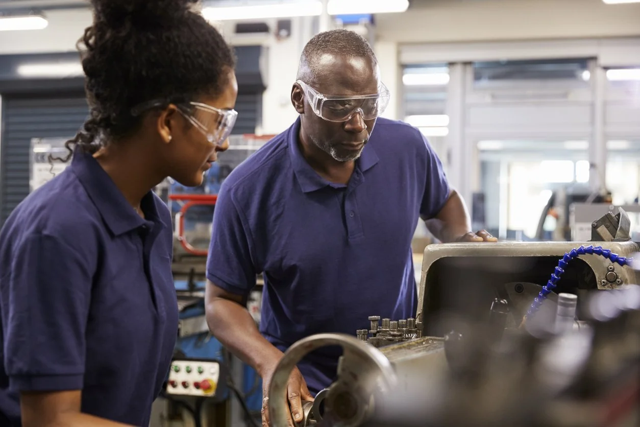 Two workers wearing safety glasses operating and inspecting machinery in a factory or workshop setting.