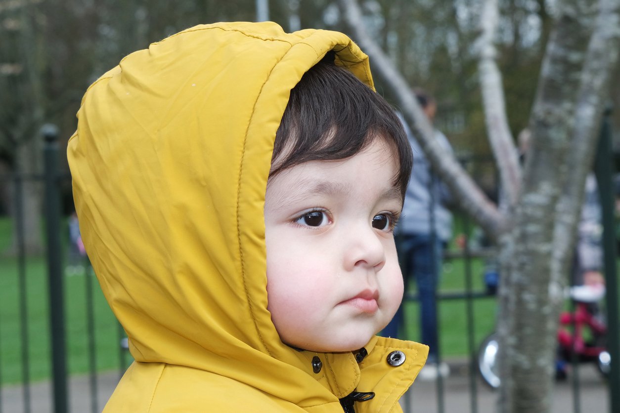Close-up of a young child with dark hair and big eyes wearing a yellow hooded jacket, outdoors near a black fence and trees.