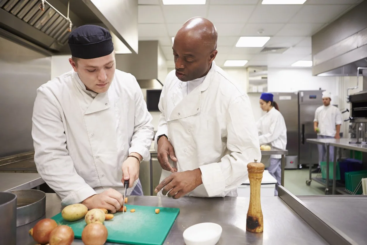 Chef teaching a young assistant how to cut vegetables in a professional kitchen, with other kitchen staff working in the background.