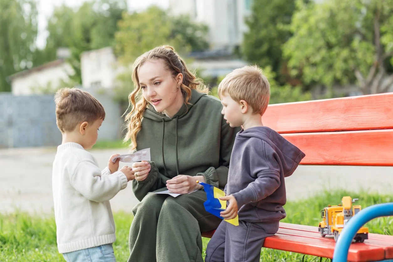 A woman with long curly hair sitting on a red park bench with two young boys, one holding a toy truck and the other showing her a paper or photo, outdoors with green trees in the background.