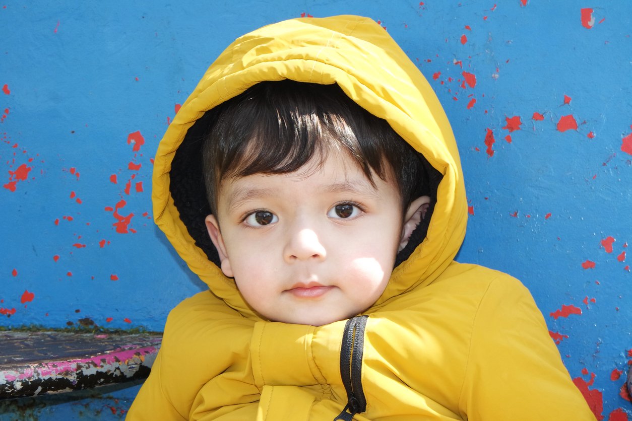 Young boy in a yellow jacket with a hood, sitting against a blue wall with red paint splatters, looking at the camera.