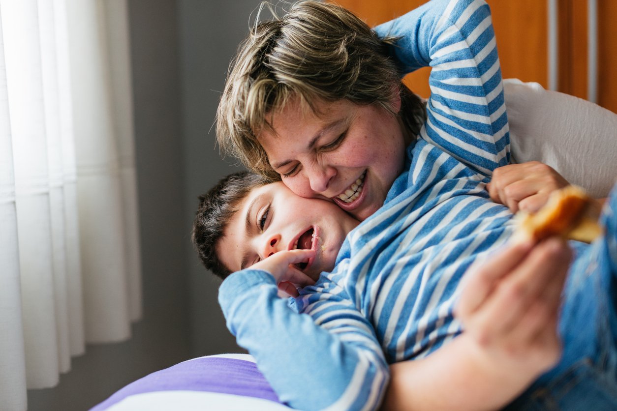 A woman and young boy playing and smiling on bed, with the woman hugging the boy with one arm and holding a piece of food in her hand.
