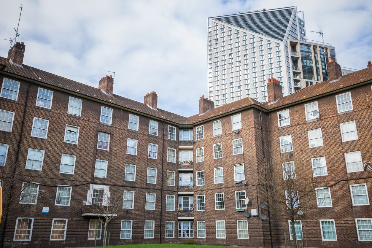 A multi-story brick apartment building with numerous windows and small trees in the foreground, and modern high-rise buildings in the background under a partly cloudy sky.
