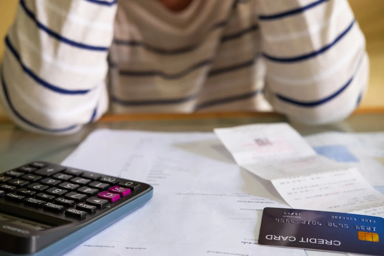 Person wearing a striped shirt sitting at a desk with a calculator, receipts, papers, and a credit card on the table.