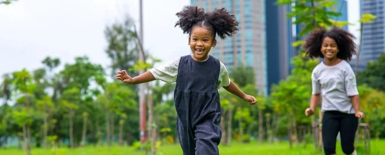 Two young girls with curly hair running and playing in a park with green trees and city buildings in the background.