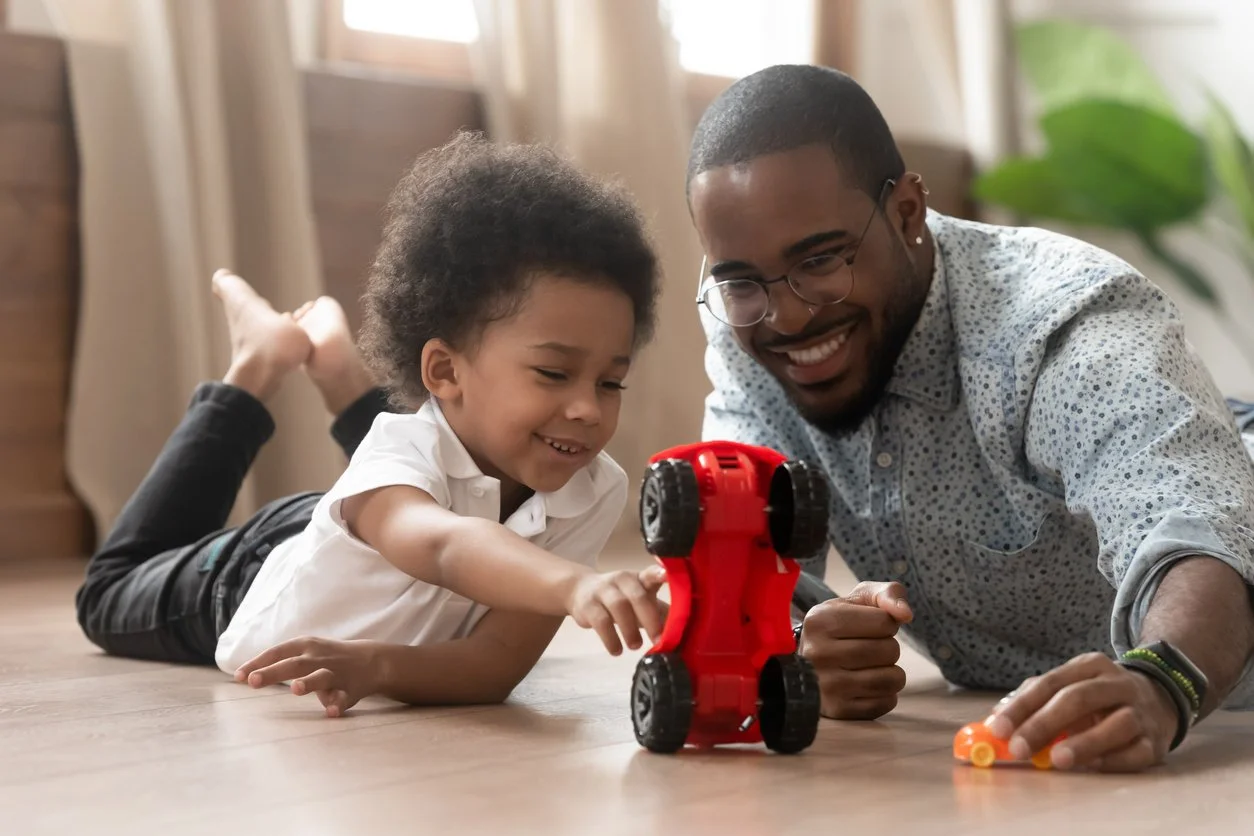 A young boy and an adult man playing with a small red toy robot on the floor in a bright indoor space.