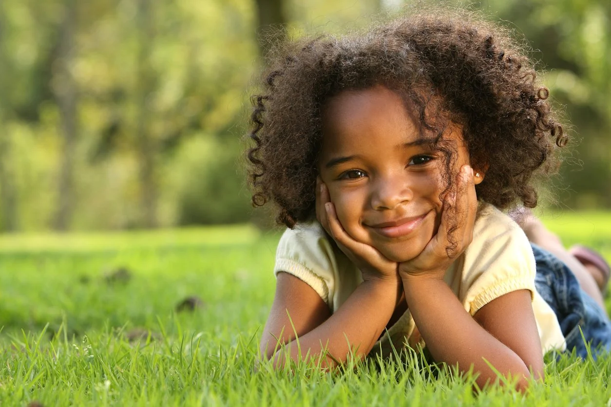 A young girl with curly hair lying on her stomach on green grass, resting her face in her hands, smiling at the camera in a park with trees in the background.