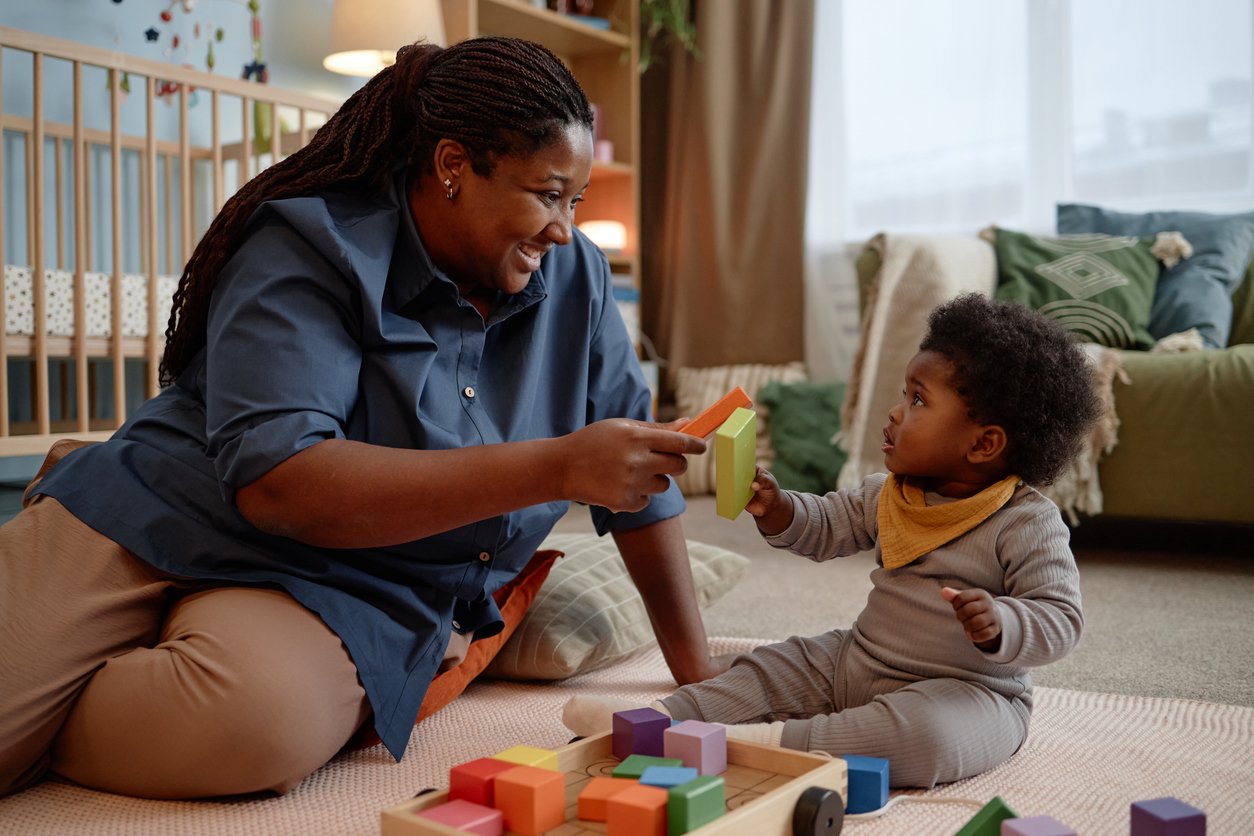 An adult woman and a young child playing with colorful wooden blocks on a carpeted floor in a cozy living room.