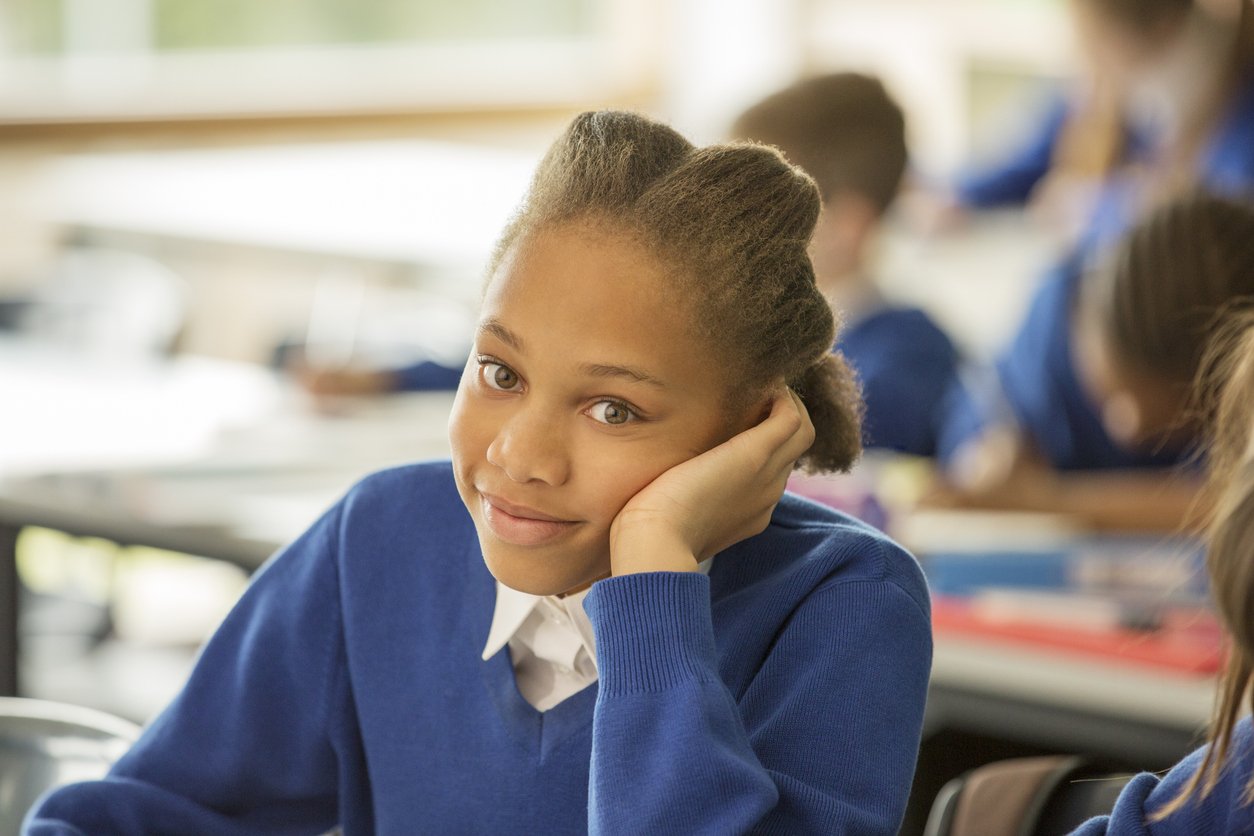 A schoolgirl in a blue uniform sitting in a classroom, resting her head on her hand, with a relaxed and slightly smiling expression, others of similar attire seated in the background.