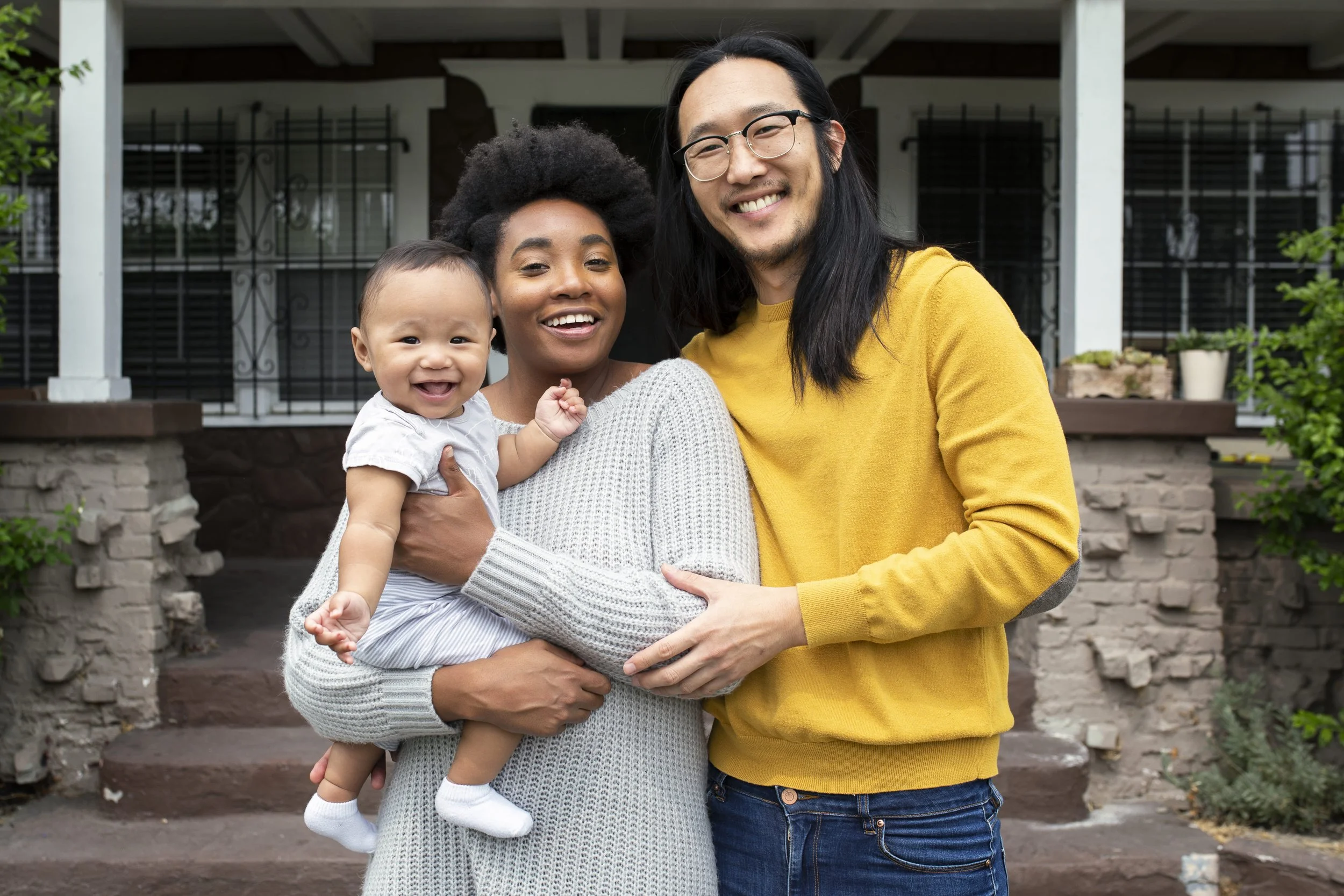 A family of three standing outside in front of a house, smiling at the camera. The woman holds a smiling baby, and the man stands next to them, wearing glasses and a yellow sweater.