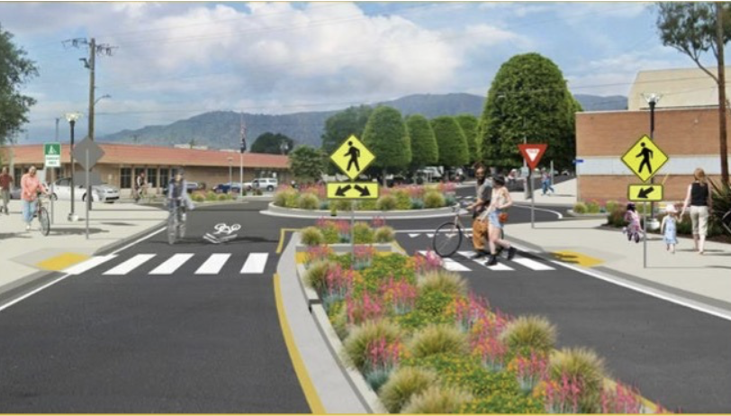 First Last Mile A crosswalk with pedestrian signals, bikeways, and traffic signs in a shopping center parking lot with a landscaped median and trees.
