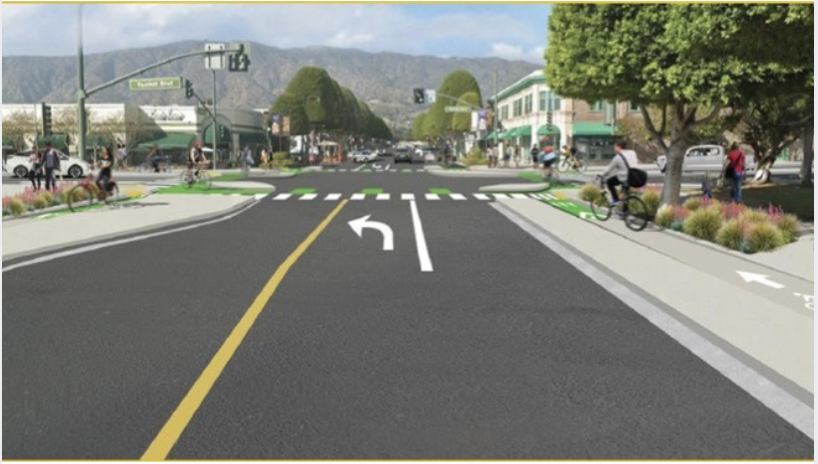 First Last Mile  A busy urban intersection with bike lanes, crosswalks, and roads for vehicles, with people riding bikes and walking, and buildings and trees in the background.
