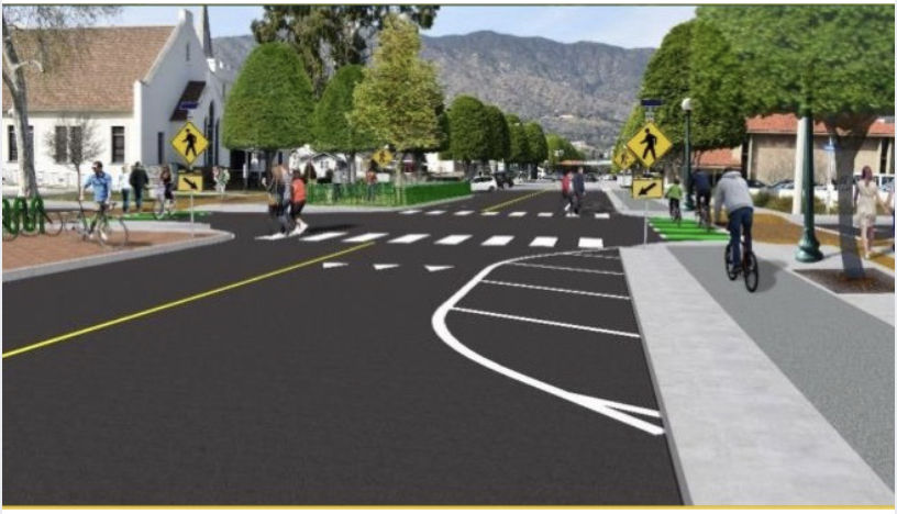 First Last Mile  A street scene with pedestrians, cyclists, crosswalks, and traffic signs in a suburban or small-town area surrounded by trees and mountains in the background.