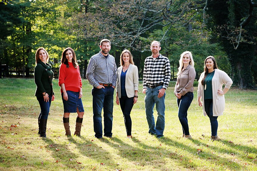 Seven people standing outdoors on a grassy area, smiling, with trees in the background.