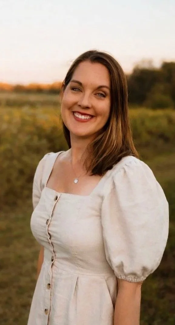 A woman with shoulder-length brown hair and light skin, smiling outdoors during sunset, wearing a beige dress with puff sleeves and buttons, standing in a field with a blurred background of grass and trees.