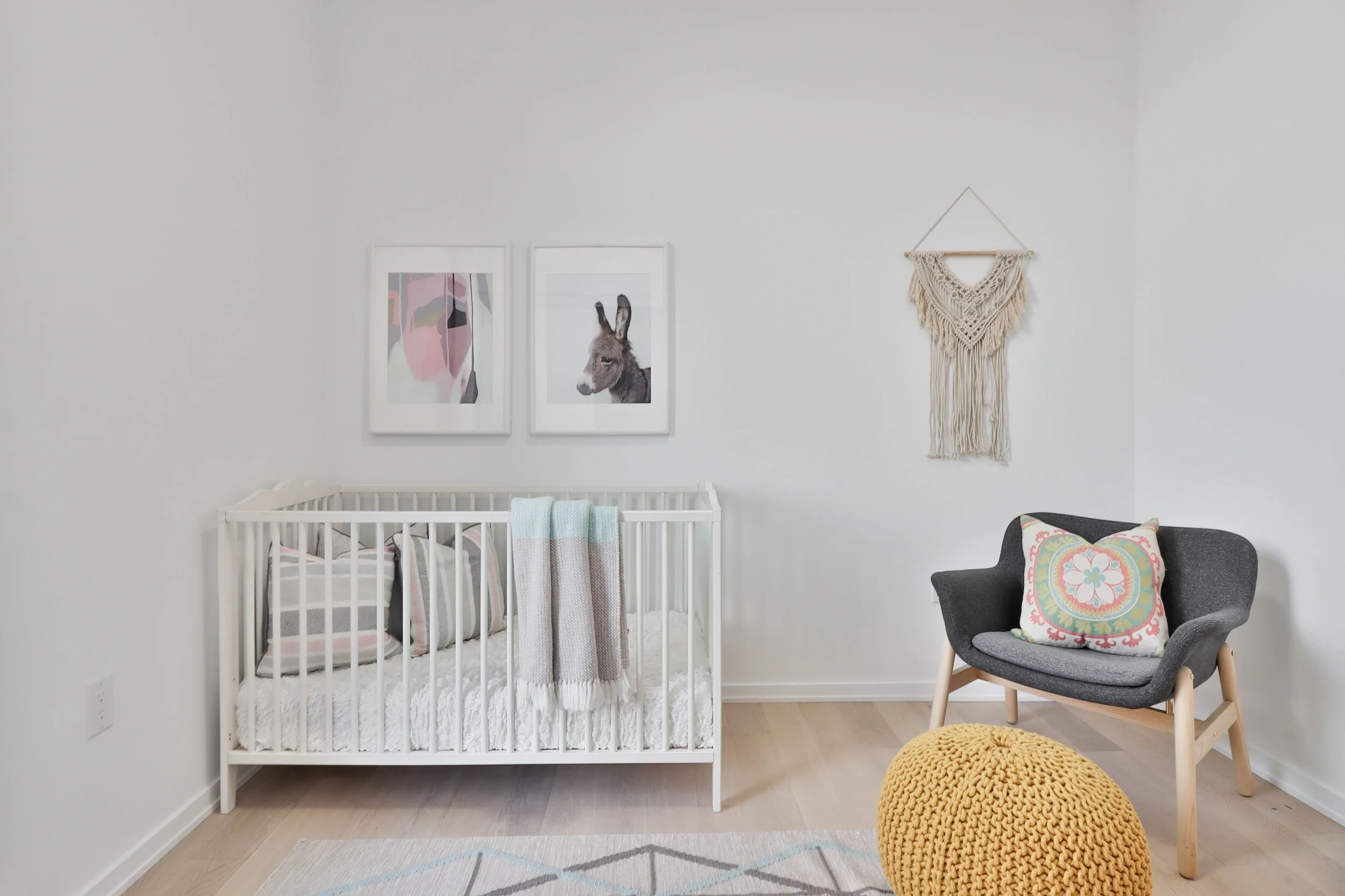 Minimalist nursery with a white crib, pastel bedding, two artworks, a gray chair with a decorative pillow, and a yellow knitted pouf.