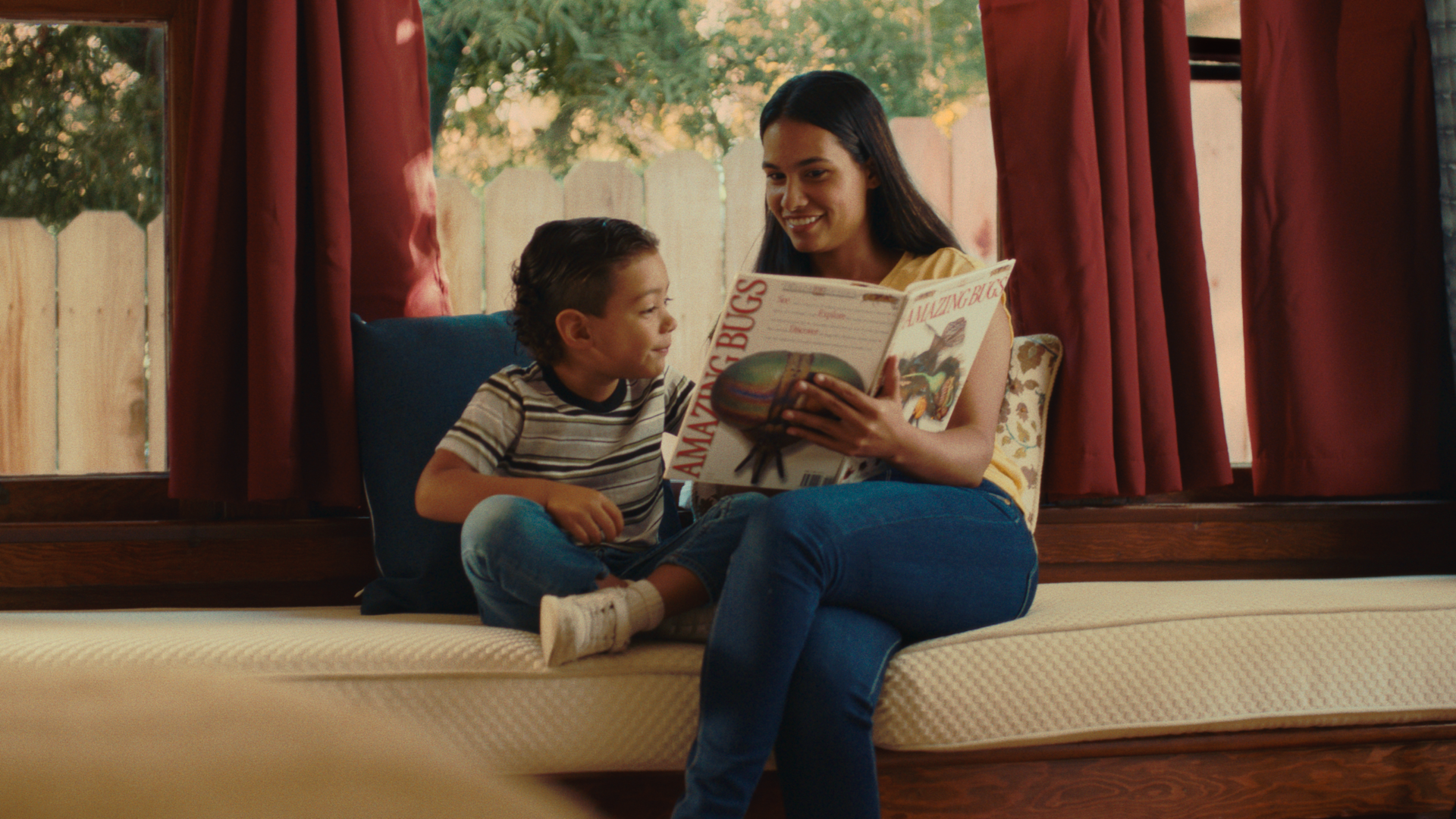 A woman and a young boy sitting on a window seat, reading a book titled 'Amazing Bugs' together, with trees visible outside the window.