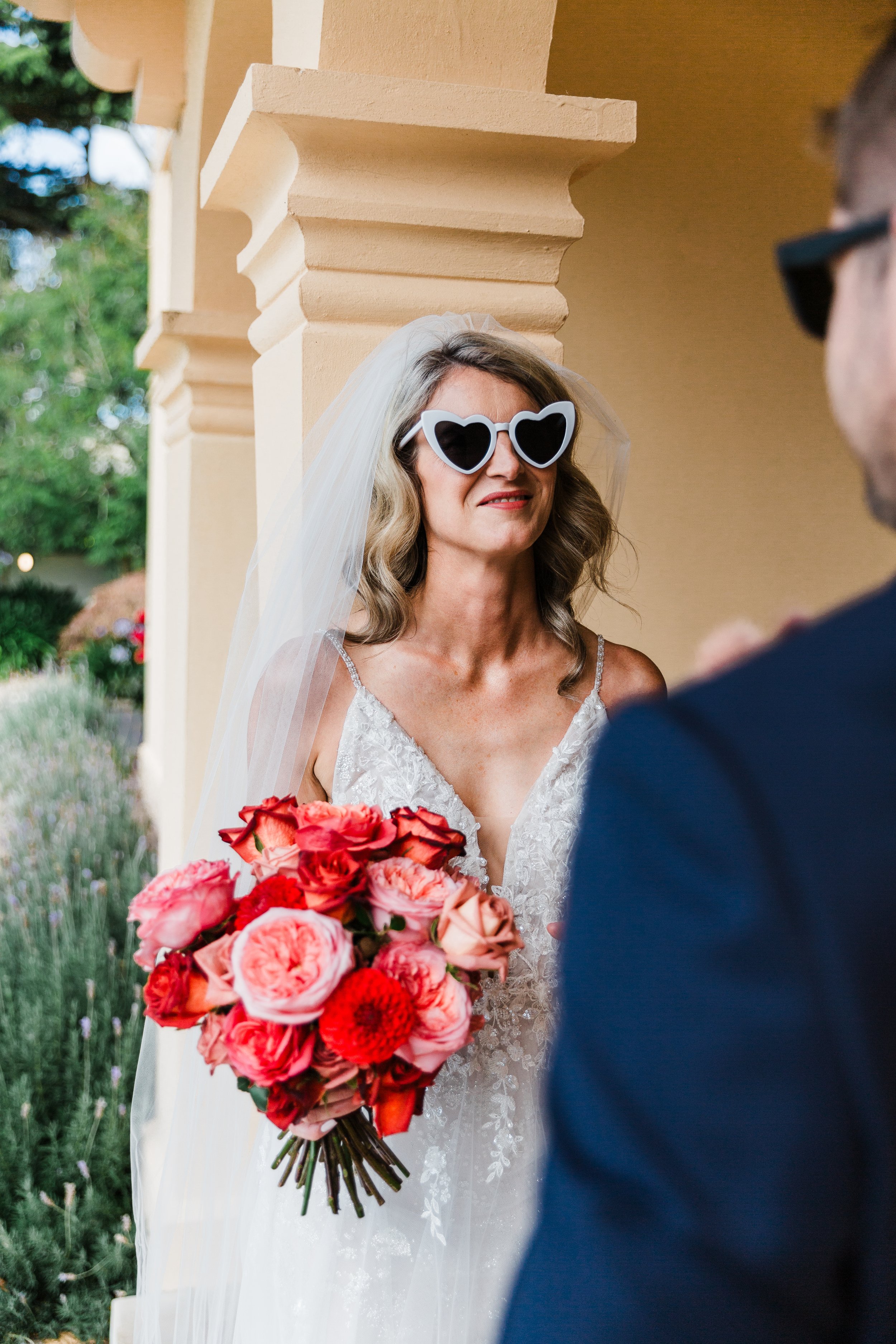 A bride wearing large white heart-shaped sunglasses and a wedding dress holding a bouquet of pink and red flowers, standing outside and smiling at a man in a suit.