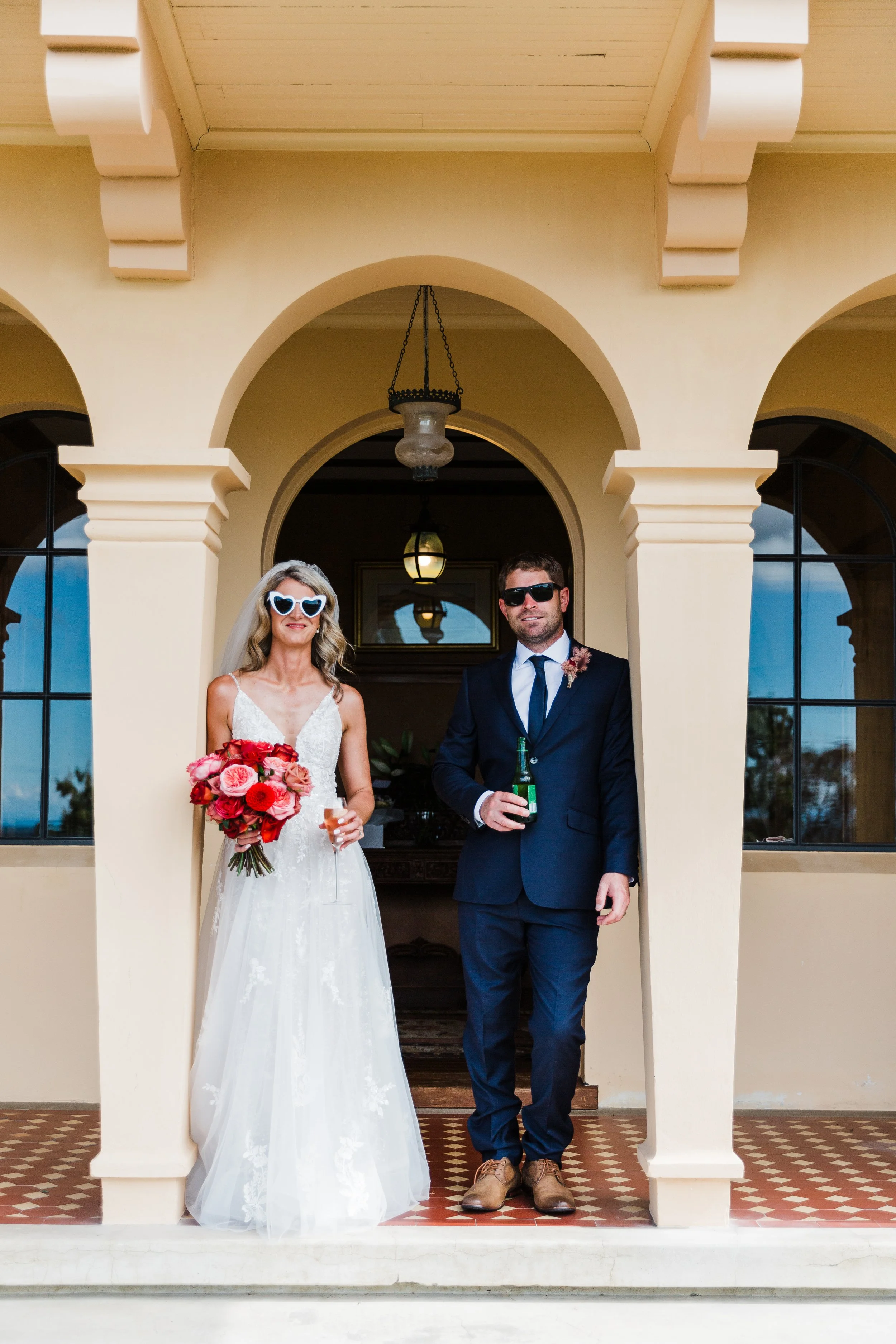 Bride and groom standing under an archway, both wearing sunglasses, holding drinks and smiling at the camera.