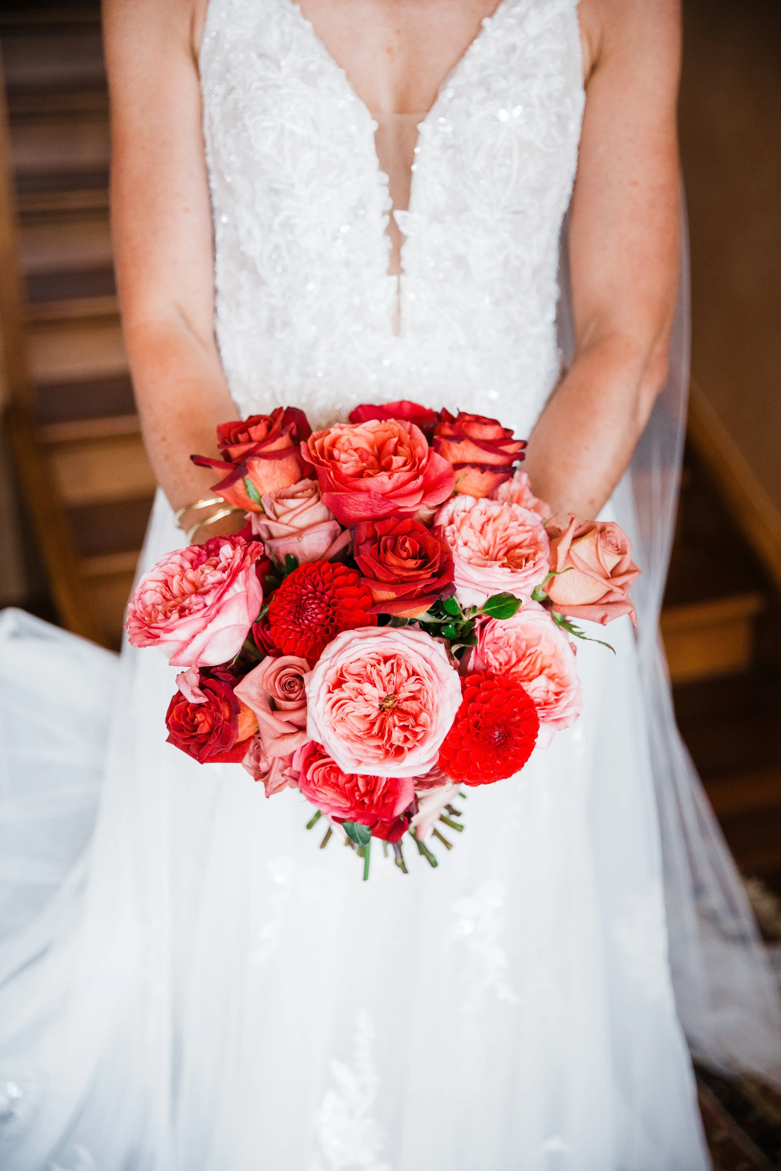 Close-up of a bride holding a vibrant bouquet of pink, red, and peach roses and dahlias, wearing a white wedding dress with lace details.