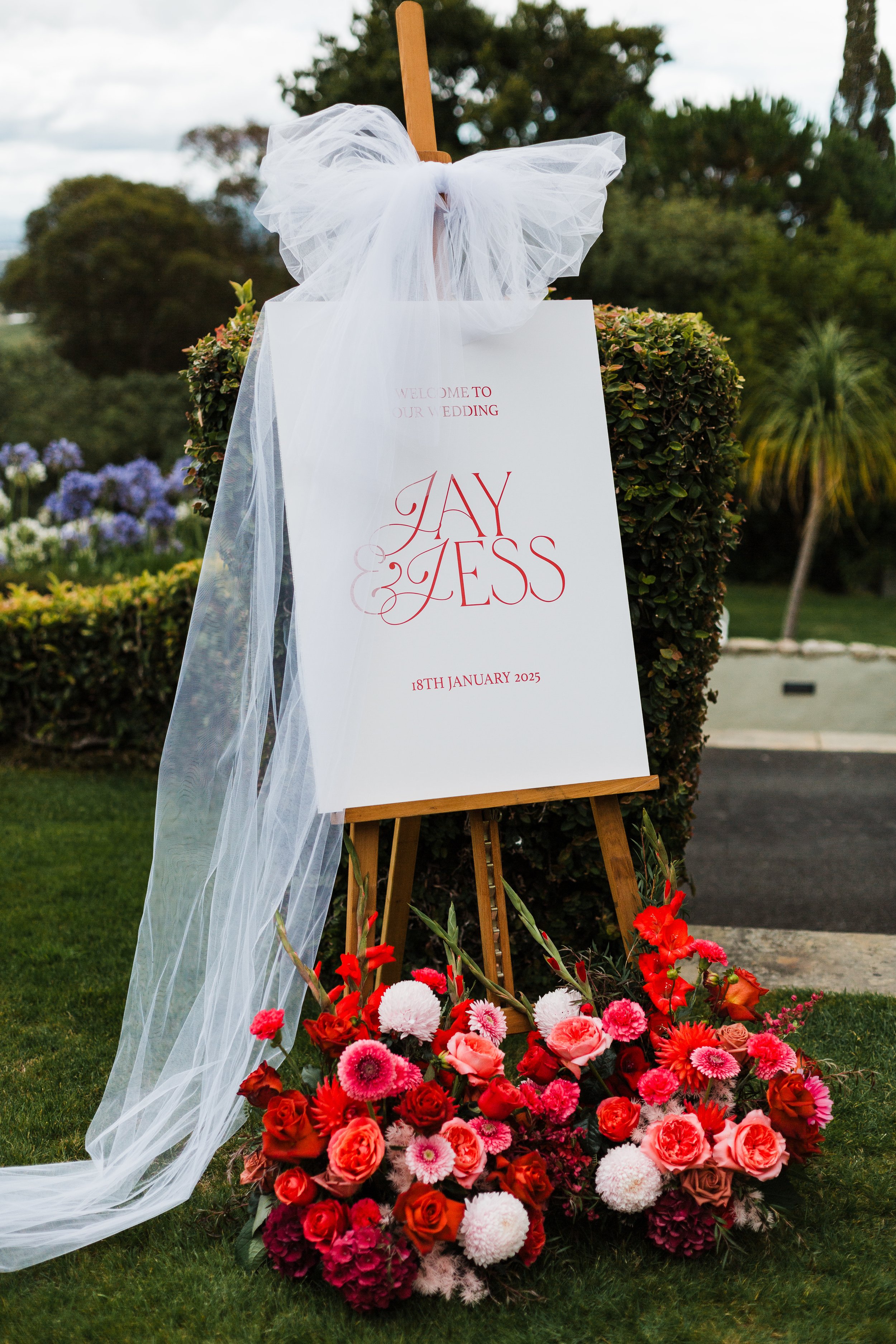 Wedding welcome sign on a wooden easel decorated with tulle, surrounded by red and pink flowers. The sign reads "Welcome to our wedding Jay & Jess, 18th January 2025." The background shows trees and a garden setting.
