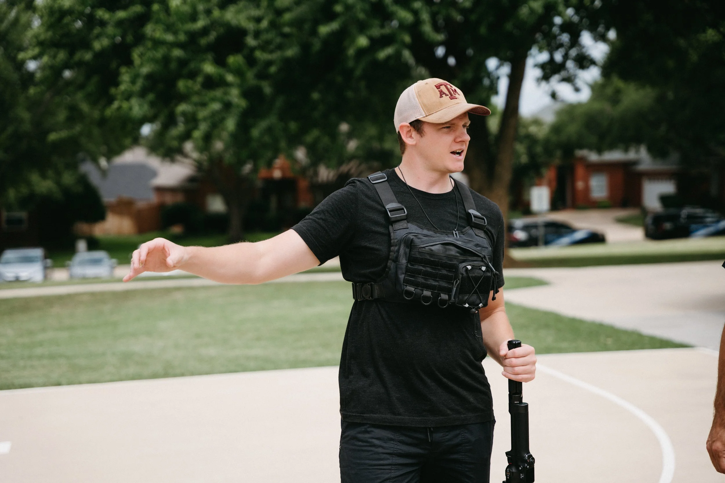 A young man wearing a khaki and red cap, black T-shirt, and black tactical vest standing outdoors on a basketball court with trees and houses in the background, holding a black tripod or monopod.