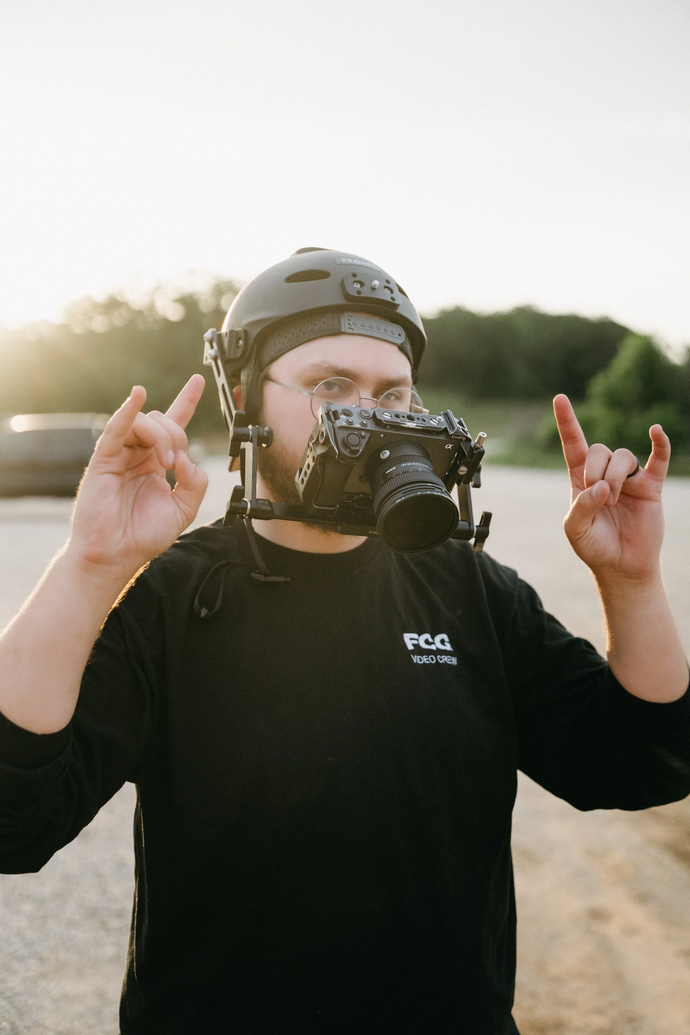 A man wearing a black helmet and glasses, holding a camera with a stabilizer rig, making a peace sign with both hands, outdoors during sunset.
