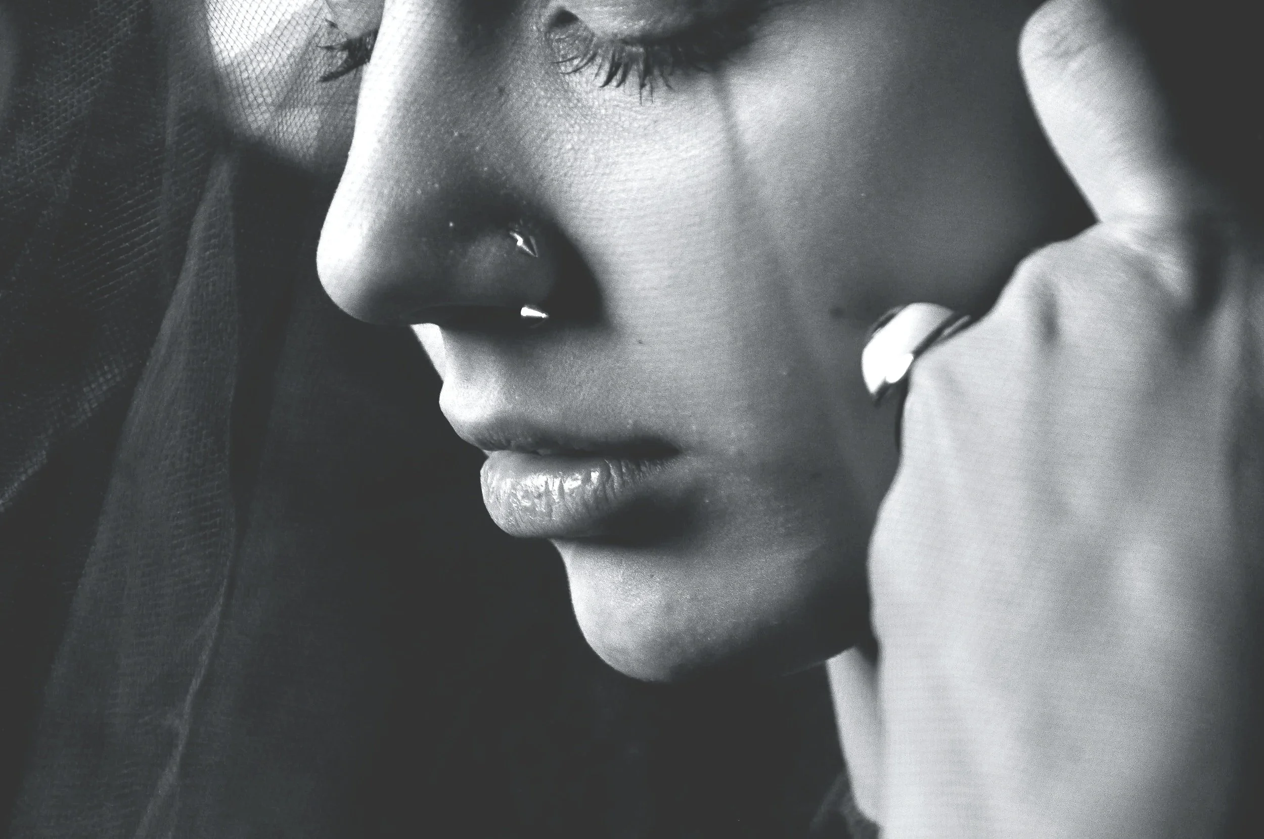 Close-up black and white photo of a woman's face, showing her nose, lips, and eye with closed eyelid, with her hand resting near her face wearing a ring.