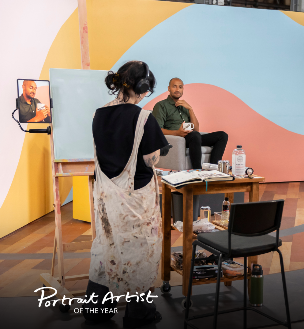 Artist standing in front of her blank canvas, preparing her palette. She is standing in front of her subject, Matt O’Kine, who is sitting on a chair in front of a colourful wall