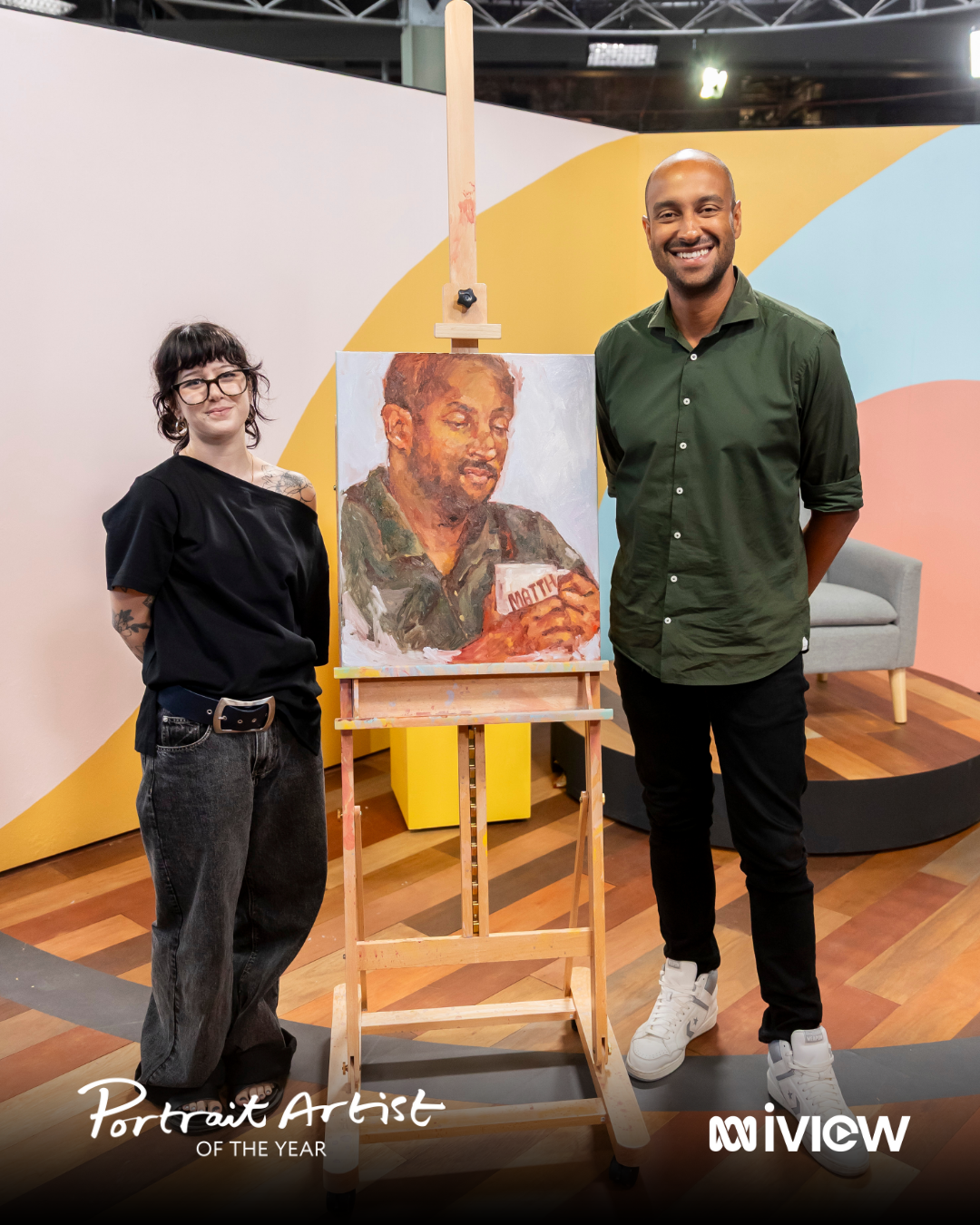 A man and a woman standing next to an easel with a portrait painting of a man holding a sign that says 'MATICS' or 'MATHE' in a studio with colorful wall and wooden floor.