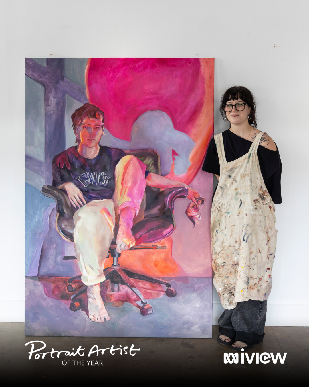 A woman with black curly hair, glasses, and tattoo on her shoulder stands next to an abstract portrait painting of a seated person with short hair, wearing a dark shirt, in front of a pink background. The woman is wearing a black top and paint-stained cream-colored apron, smiling at the camera.