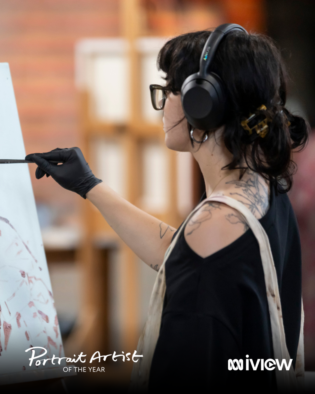 A woman with dark hair, glasses, tattoos, and wearing black gloves is painting on a canvas while wearing large headphones.