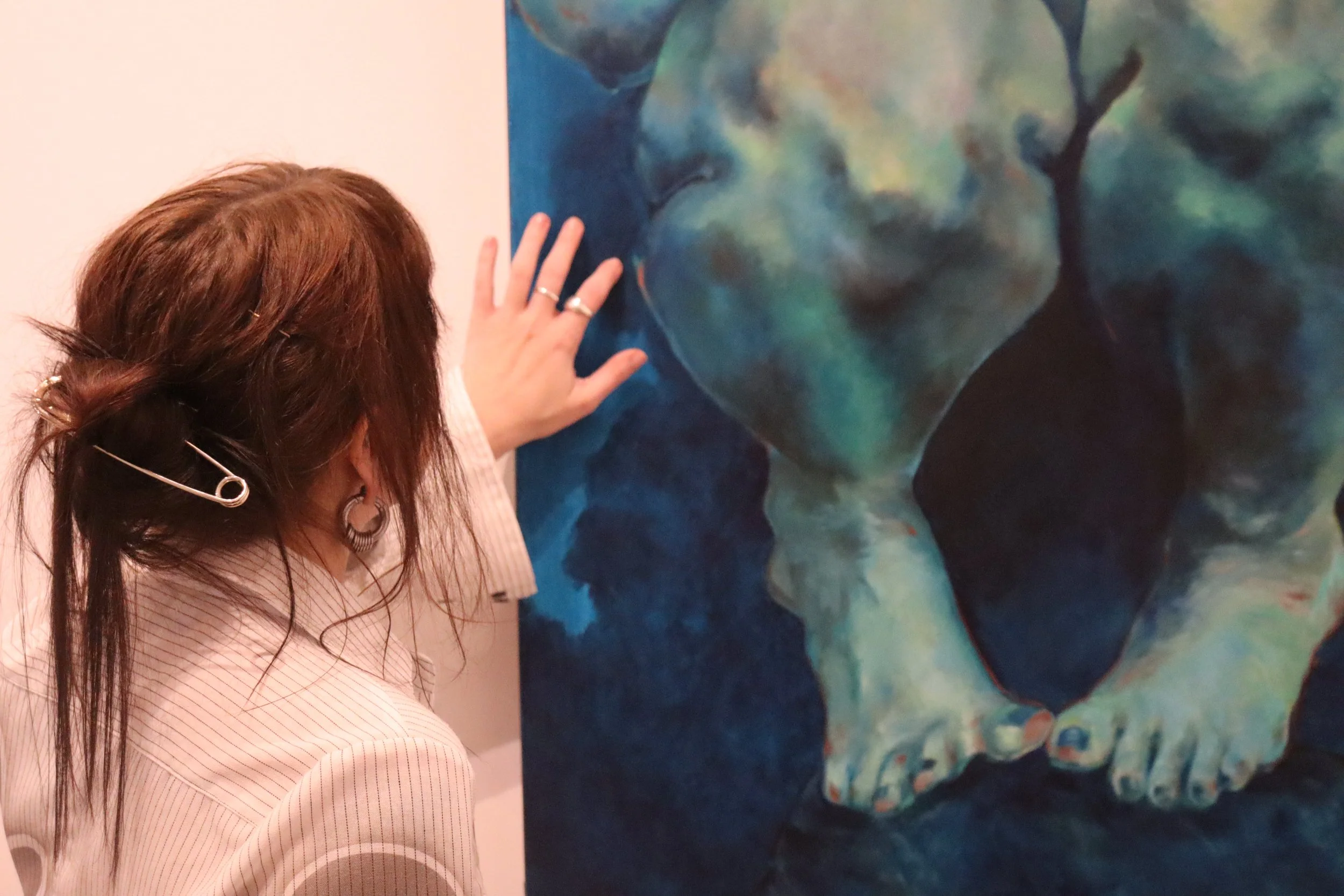 A woman with long brown hair and rings on her fingers is touching a large abstract painting that features shades of blue and green.