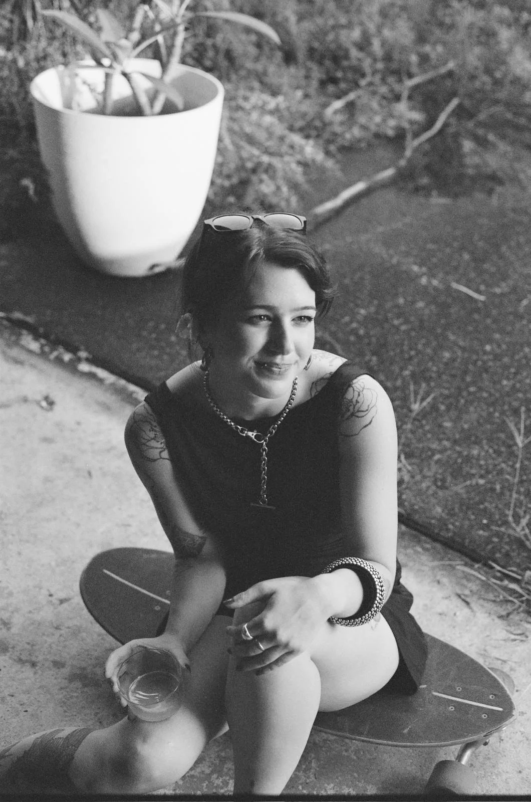 A woman with short dark hair, tattoos, and a necklace, sitting on a skateboard outdoors, holding a glass of drink, with sunglasses on her head and a smile.