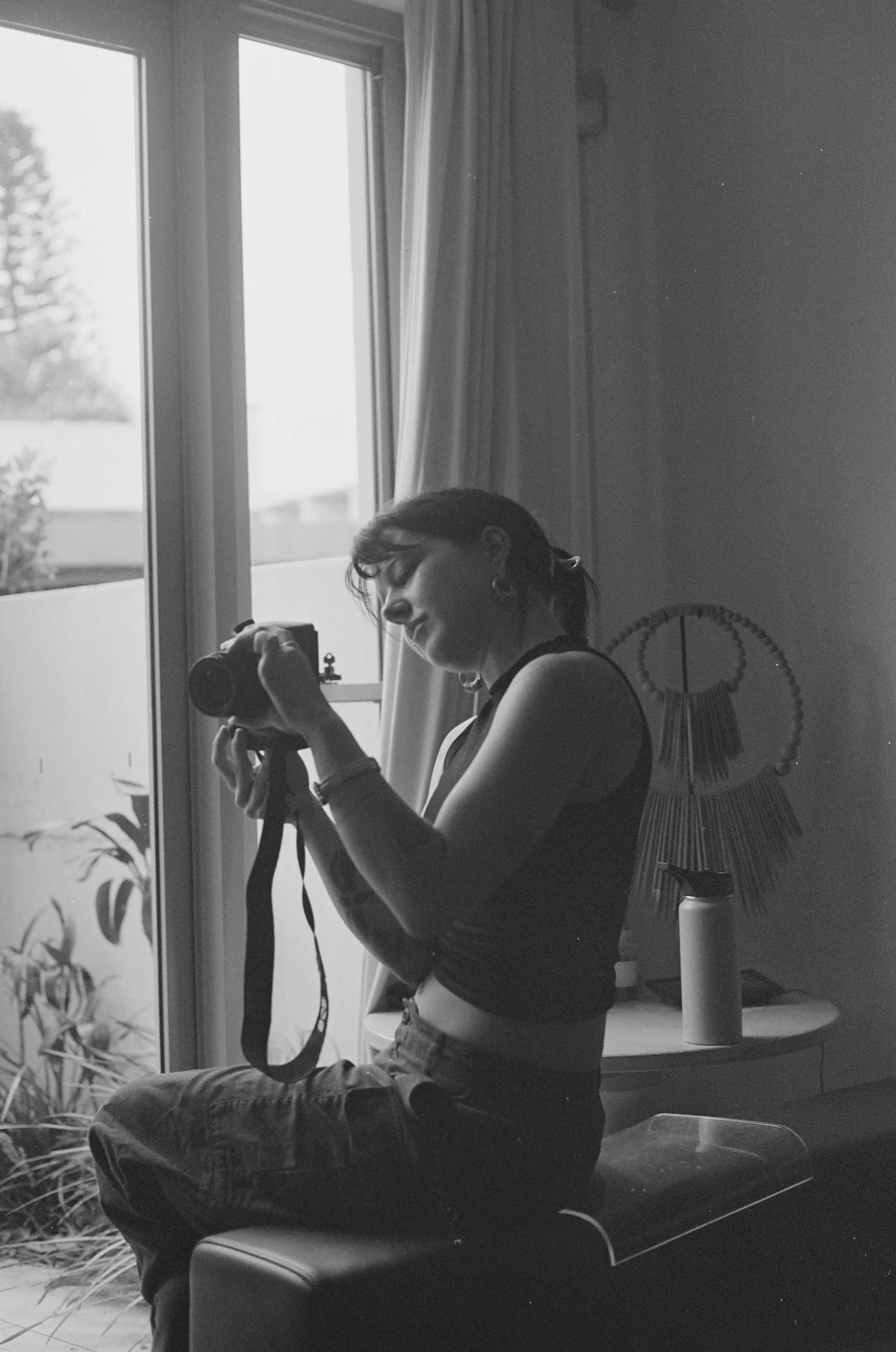 A woman sitting on a bench, taking a photo with her camera, near a window with curtains, with a lamp and some wall decor in a home interior, illuminated by natural light.