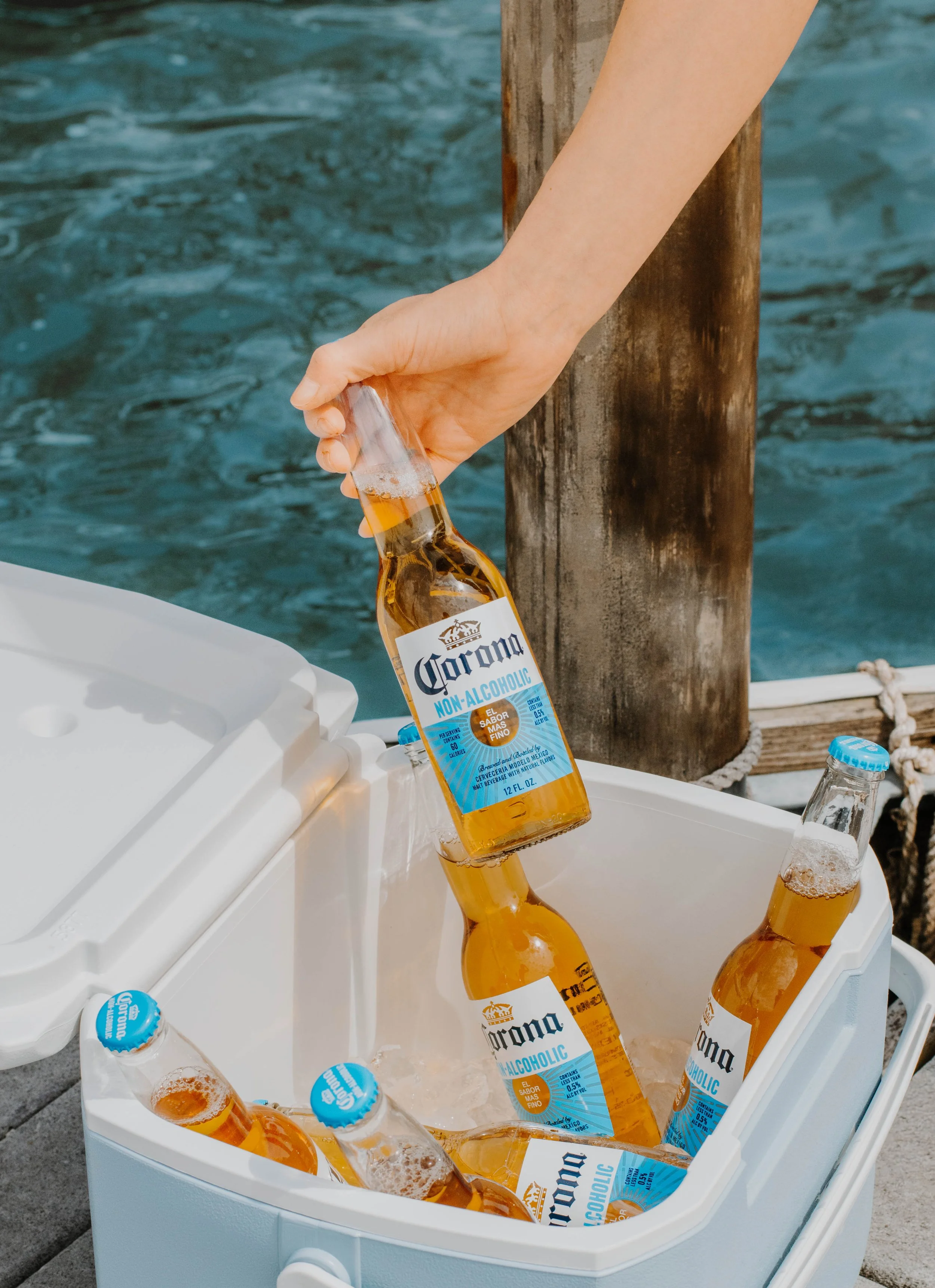 Hand placing a Corona non-alcoholic beer bottle into a white cooler, with other bottles and water bottles inside, near a wooden post and water background.