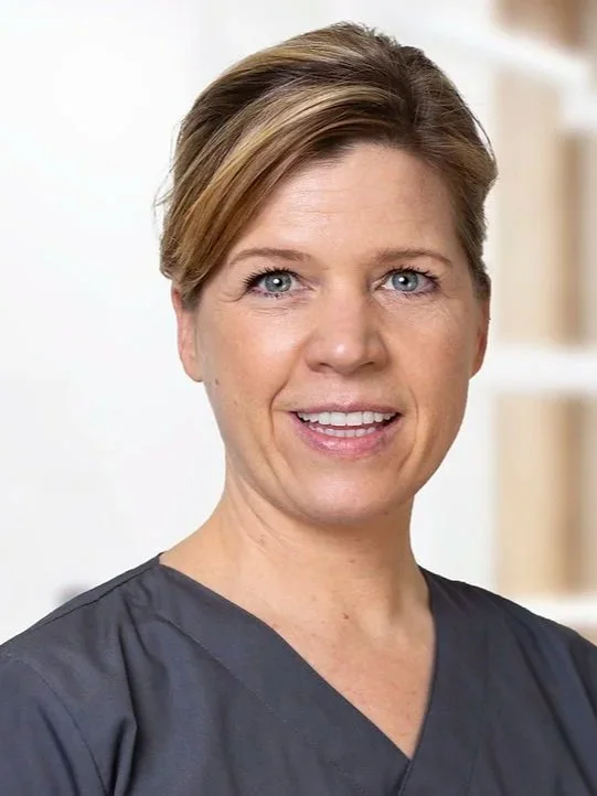Nahaufnahme einer Frau mit blauen Augen und kurzen, hellbraunen Haaren, die eine graue medizinische Uniform trägt, vor einem neutralen Hintergrund.