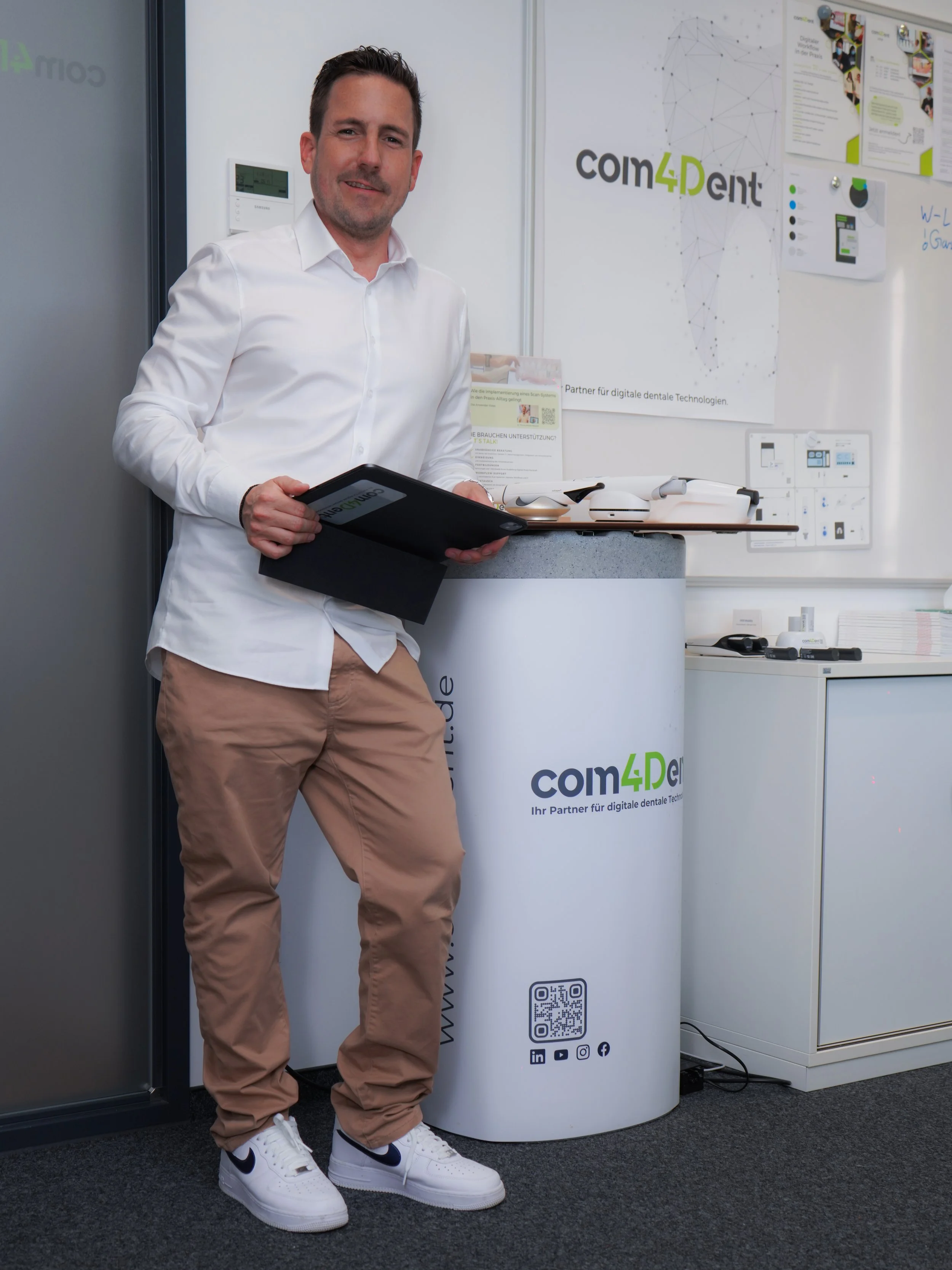 Fabian Lorenz Geschäftsführer com4Dent steht vor einem Whiteboard mit Plakaten und einem Banner, das 'com4dent' heißt, und hält ein Tablet in der Hand. Die Umgebung sieht nach einem Büro oder einer Präsentationsfläche aus. in Bad Kreuznach