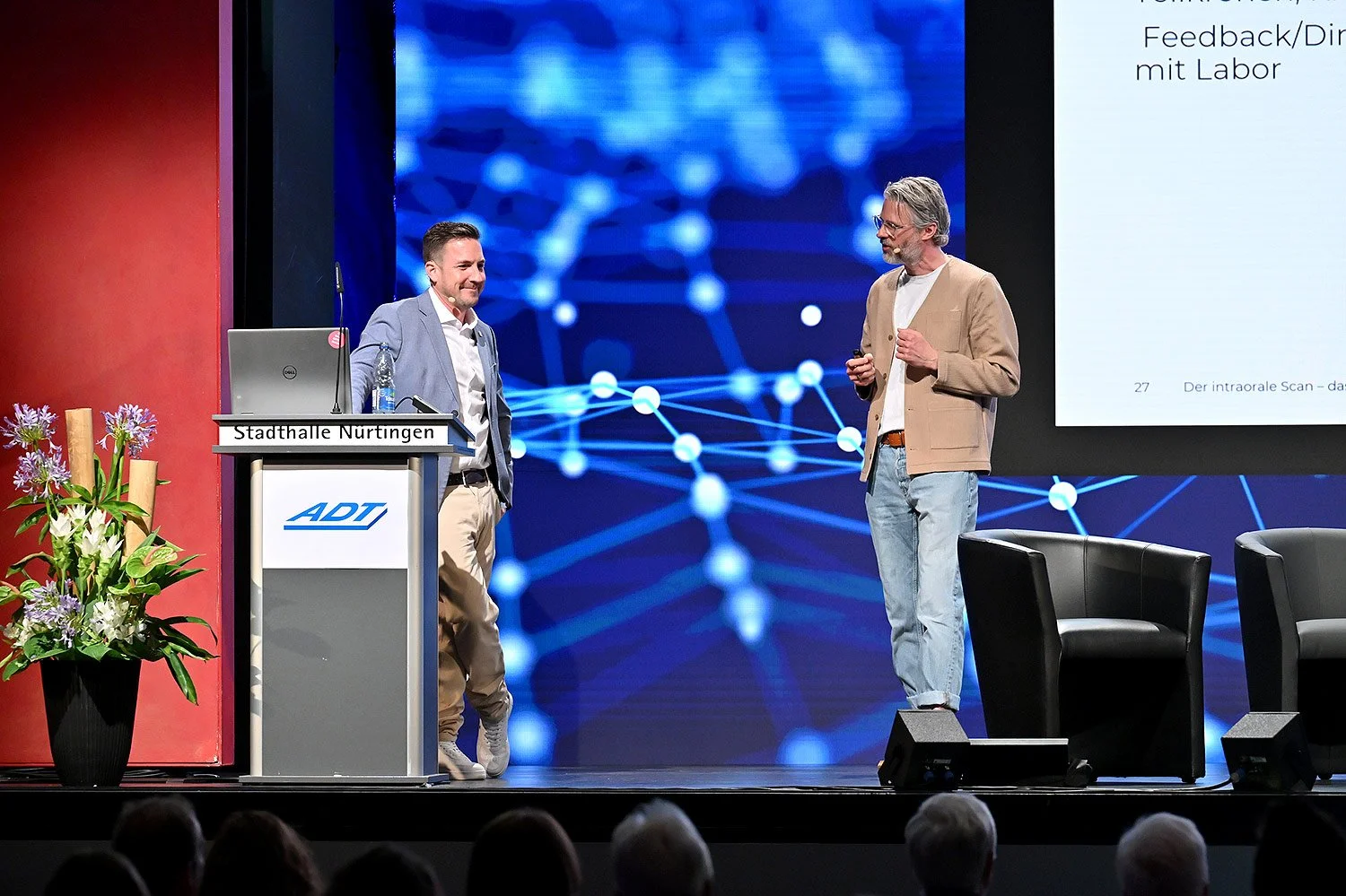 Fabian Lorenz bei dem ADT-Kongress in Nürtingen Einer steht hinter einem Podium mit dem Logo 'ADT' und einem Laptop, der andere spricht. Im Hintergrund ist eine große Leinwand mit einem digitalen Netzwerk-Hintergrund.