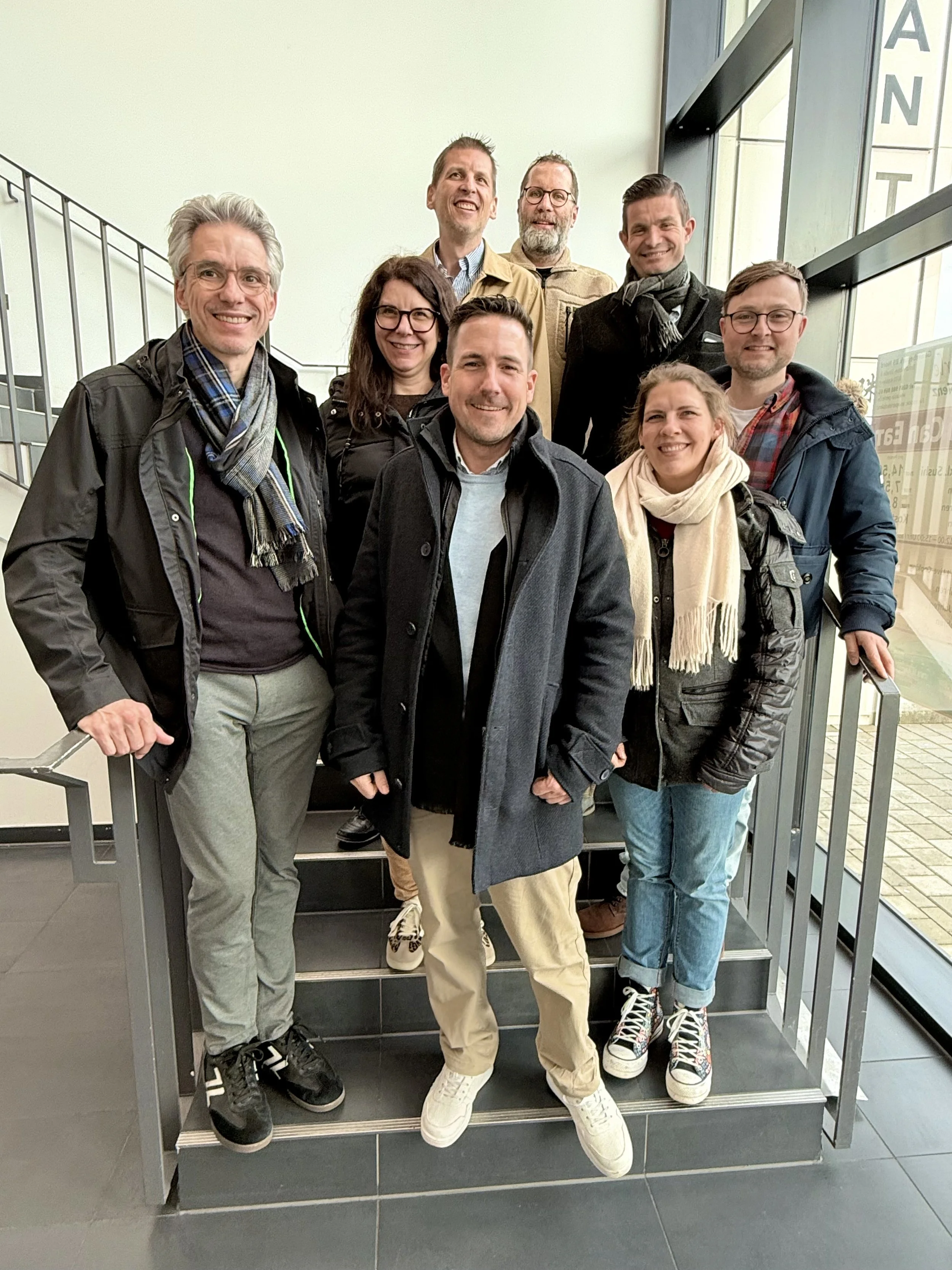 Eine Gruppe von acht Menschen steht auf einer Treppe in einem Gebäude und lächelt.