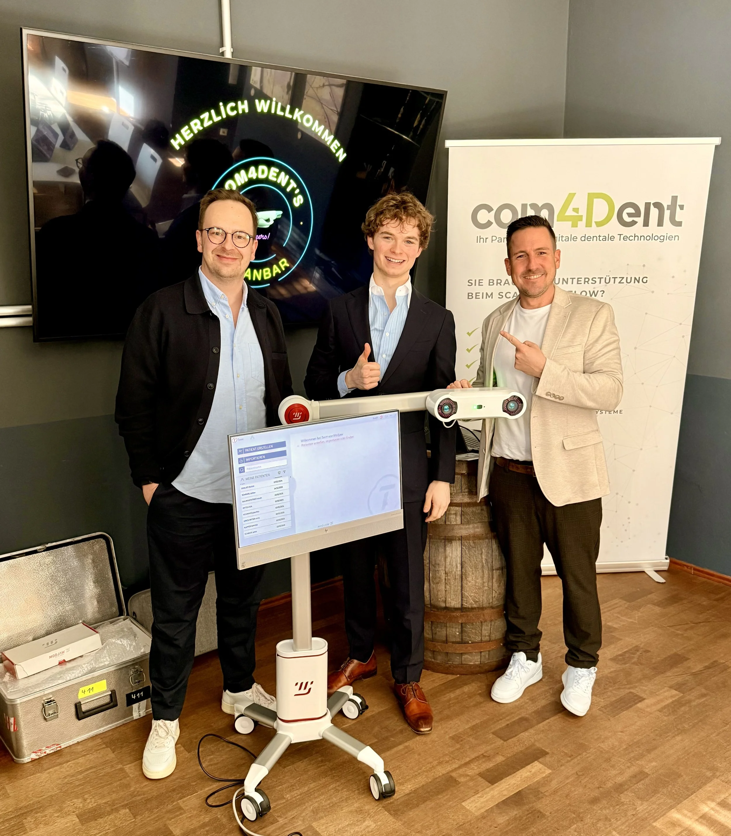 Drei Männer stehen neben einem weißen, fahrbaren Robotertisch auf Holzfußboden. Im Hintergrund sind ein Bildschirm mit Begrüßungstext und eine Werbetafel mit dem Logo 'com4Dent'. Die Männer lächeln freundlich, eine Person macht ein Daumen-hoch-Symbol, eine andere zeigt auf den Roboter.